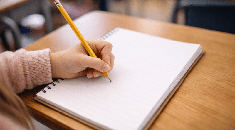 Child's hand holding a pencil, writing in a spiral-bound notebook.